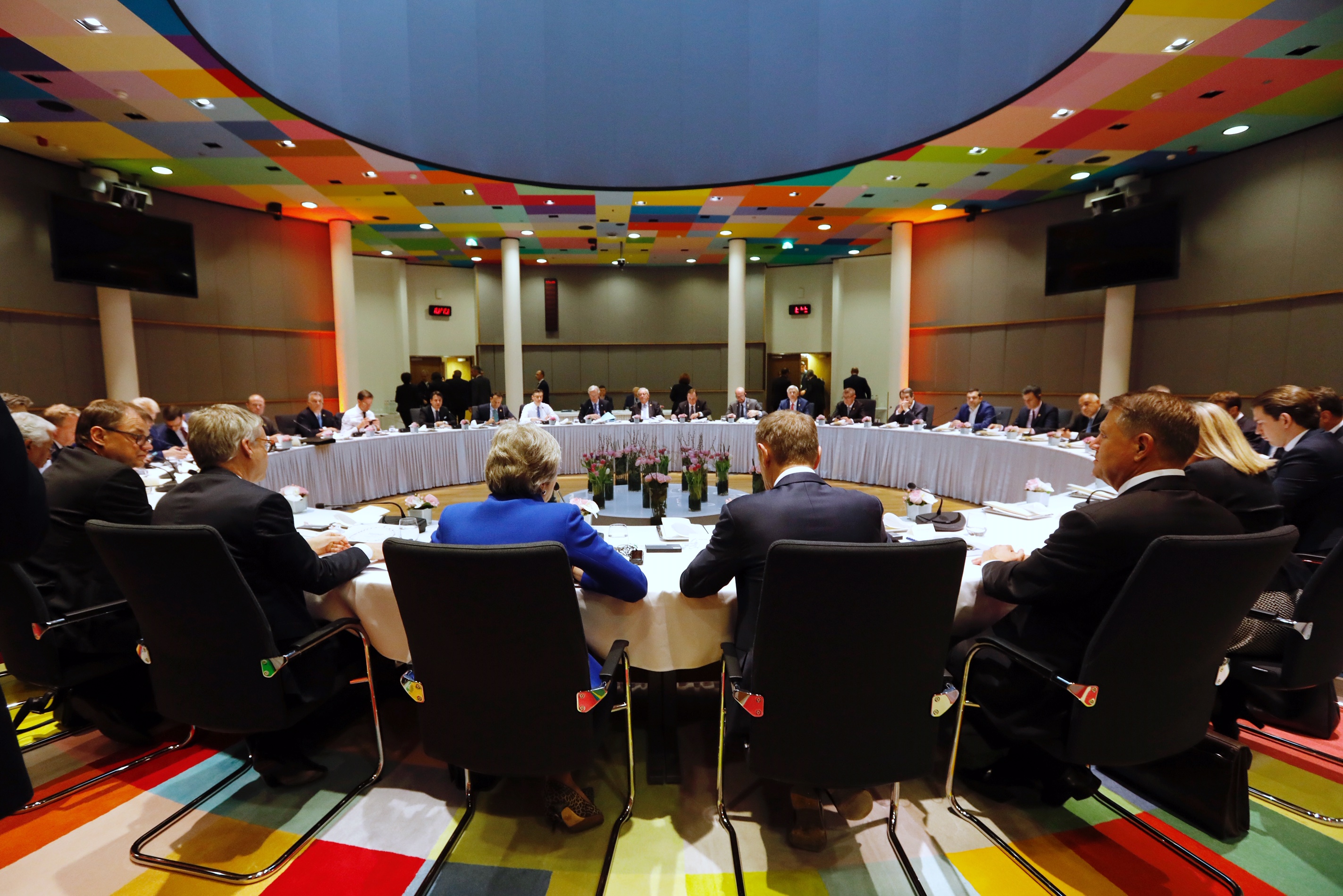 Duży okrągły stół przy którym siedzą szefowie państw lub rządów Unii Europejskiej. Na pierwszym planie tyłem Donald Tusk i Teresa May. Kolorowa podłoga i sufit.