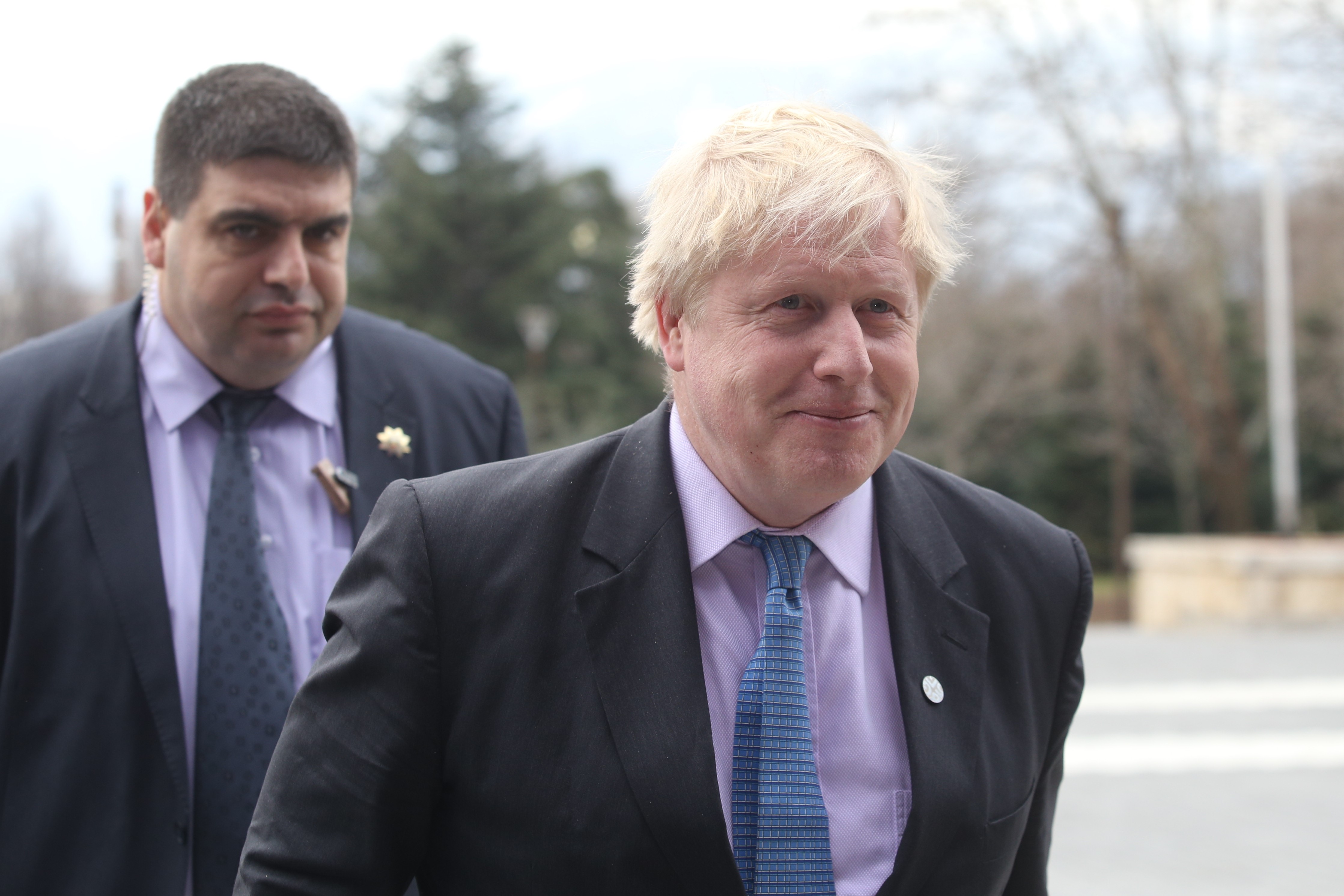Premier Wielkiej Brytanii Boris Johnson. Zdjęcie przedstawia premiera na dworze, ujęcie przedstawia ramiona oraz głowę. Premier ubrany jest w granatową marynarkę, fioletową koszulę oraz niebieski krawat. W klapie ma srebrną wpinkę. Za premierem kroczy ochroniarz. W tle widać drzewa.