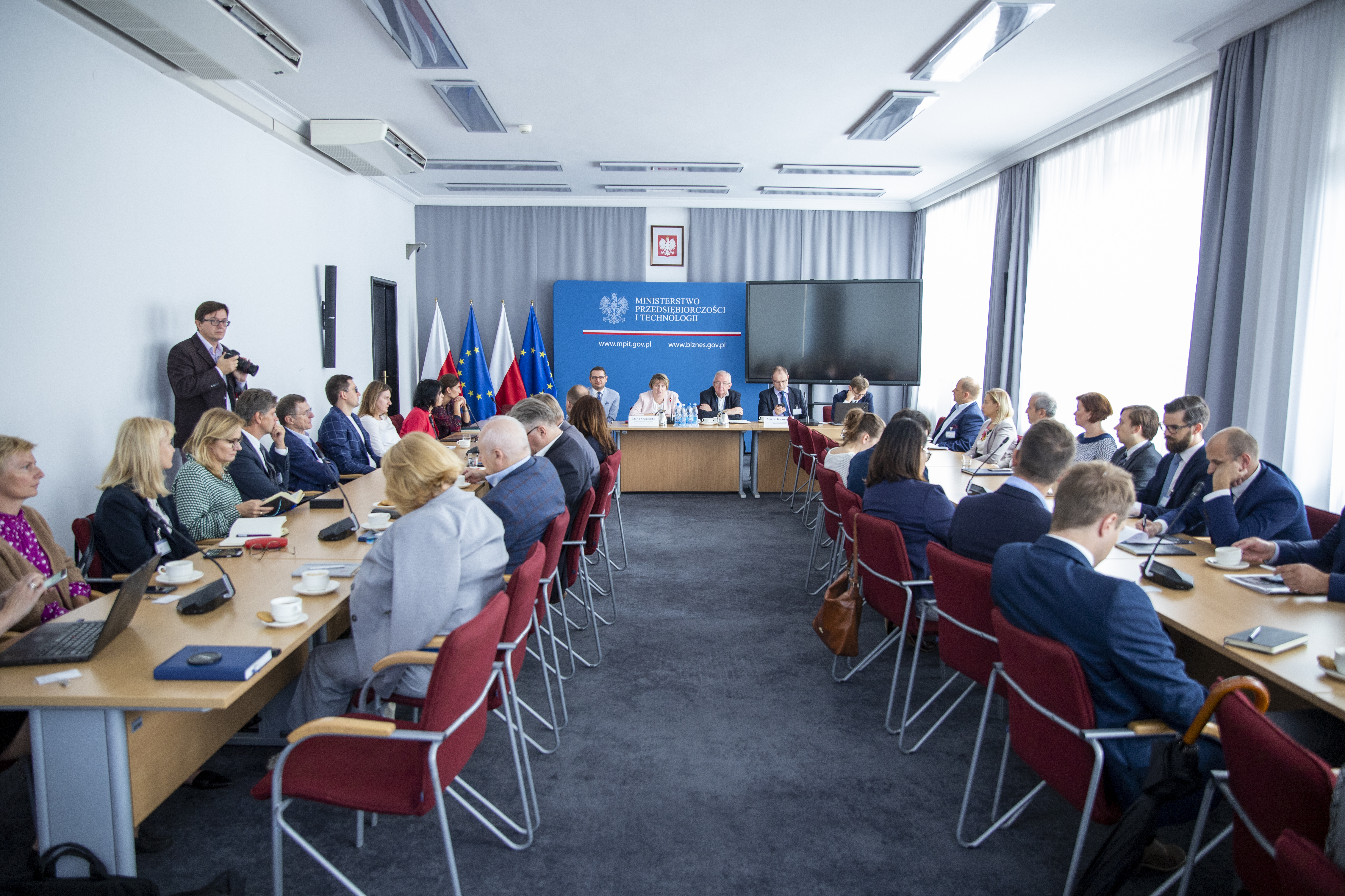 Zdjęcie przedstawia uczestników spotkania siedzących przy dwóch stołach oraz prelegentów na tle niebieskiej tablicy Ministerstwa Przedsiębiorczości i Technologii.