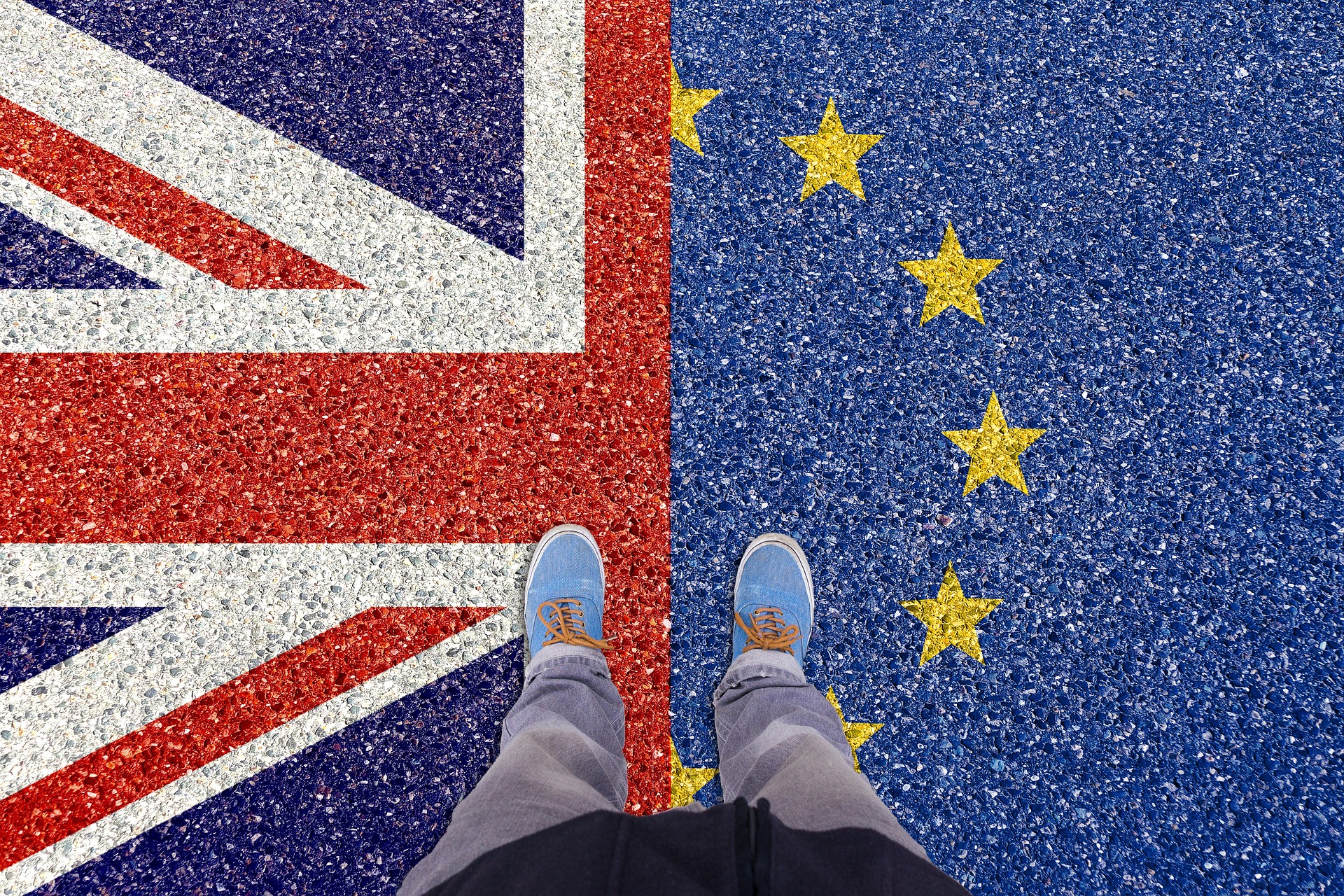 Obraz przestawia buty człowieka stojącego na ziemi na której nadrukowana jest flaga UE i Wielkiej Brytanii