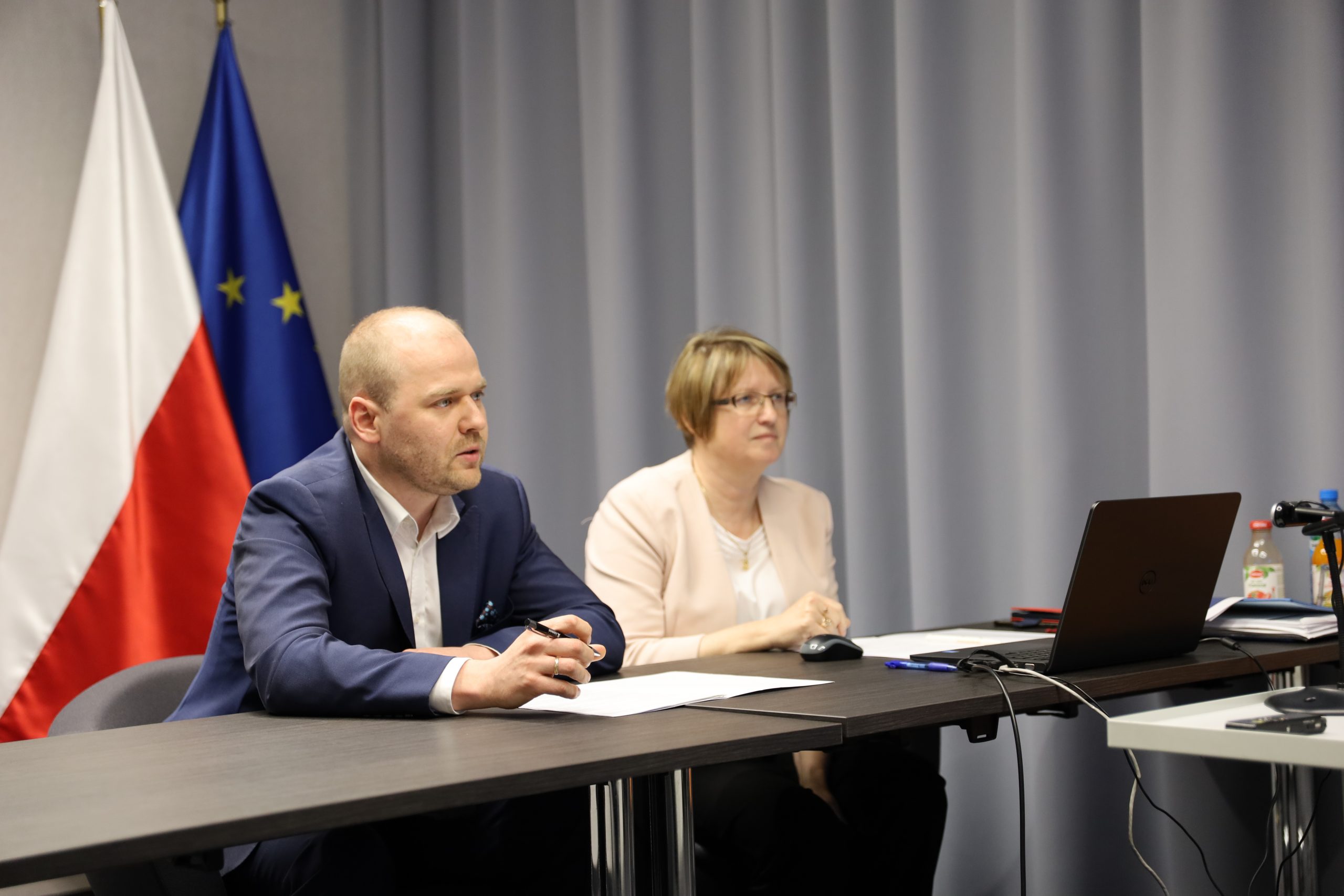 Zdjęcie przedstawia Ministra Krzysztofa Mazura oraz Panią Dyrektor Marię Szymańską. Oboje siedzą za stołem. W tyle widać flagi Polski oraz Unii Europejskiej.